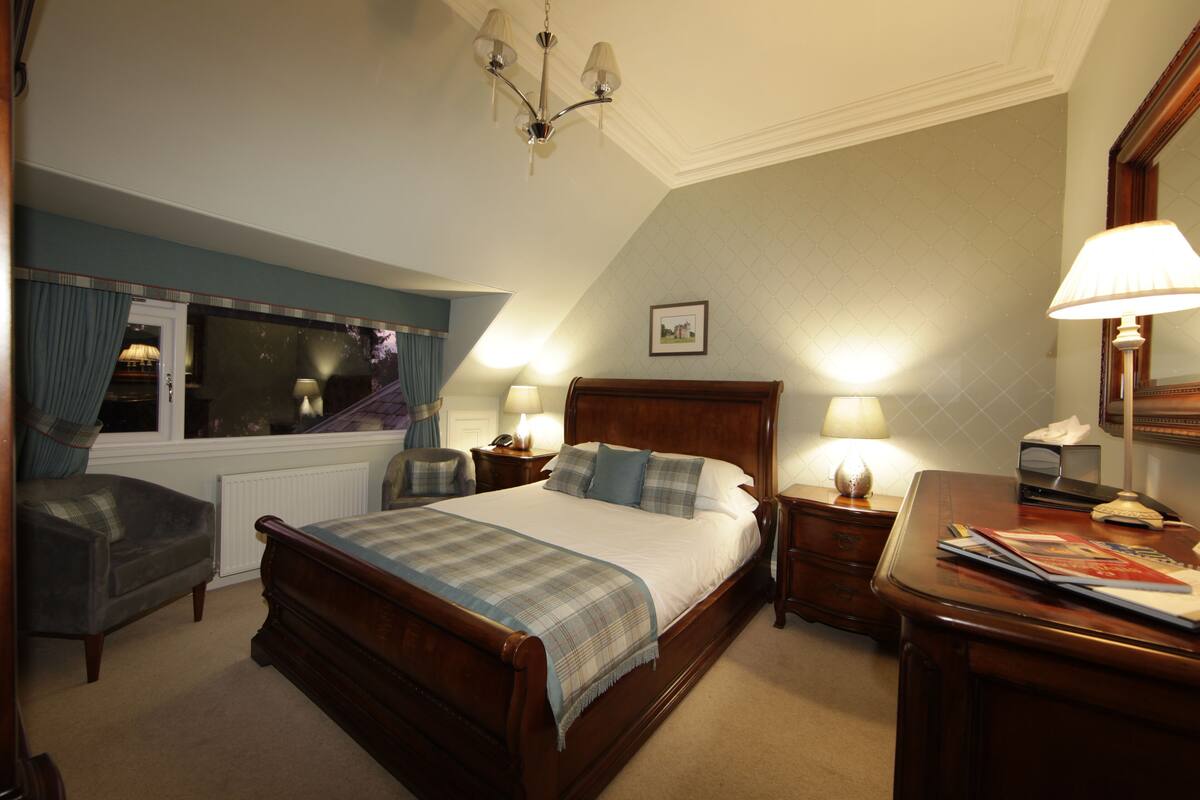 A bedroom with a large bed, a wooden nightstand, a chair, a desk, and a window with curtains.