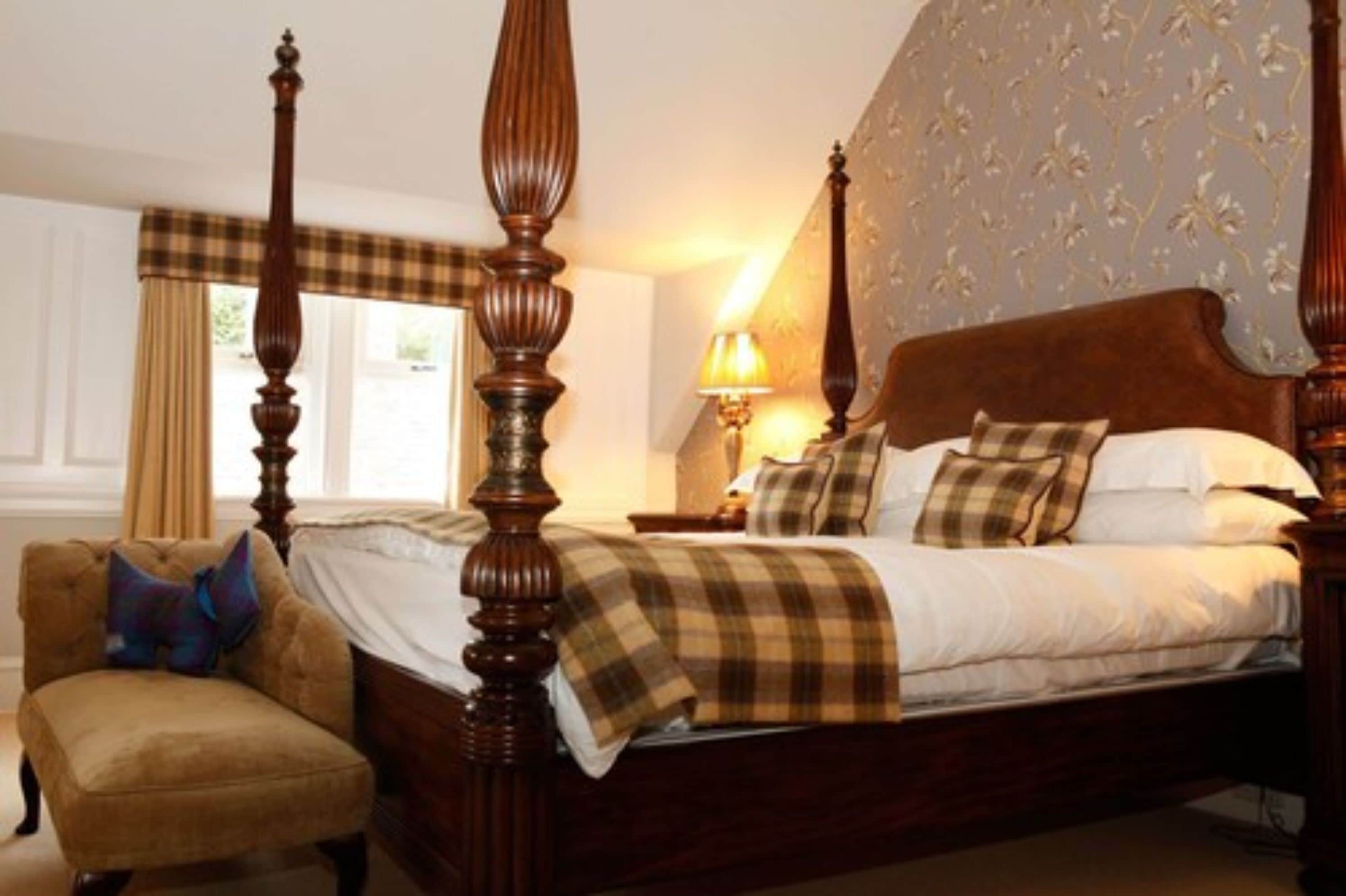 Una cama con dosel y tablero de madera, una mesita de noche con lámpara, una silla y una ventana con cortinas.
