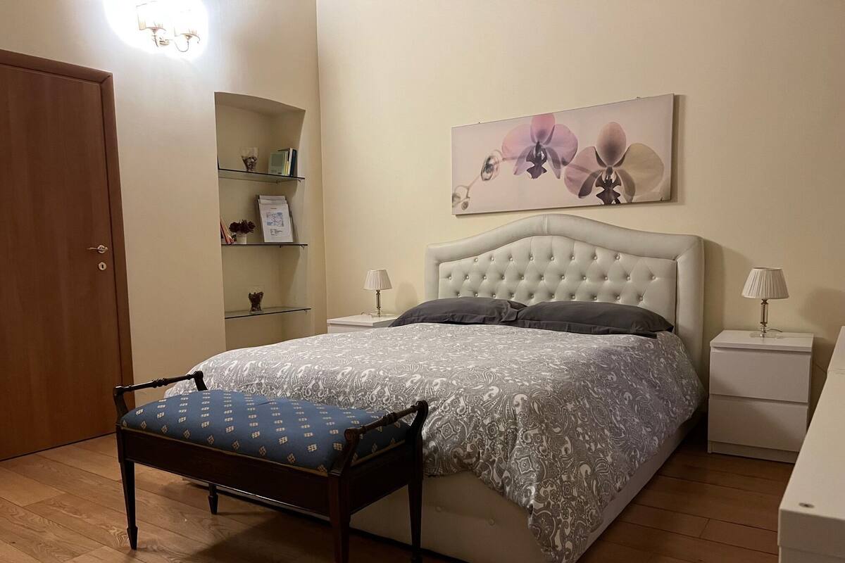 A bedroom with a bed, bedside tables, a chair, a shelf with books and decorative items, and a wall-mounted picture.