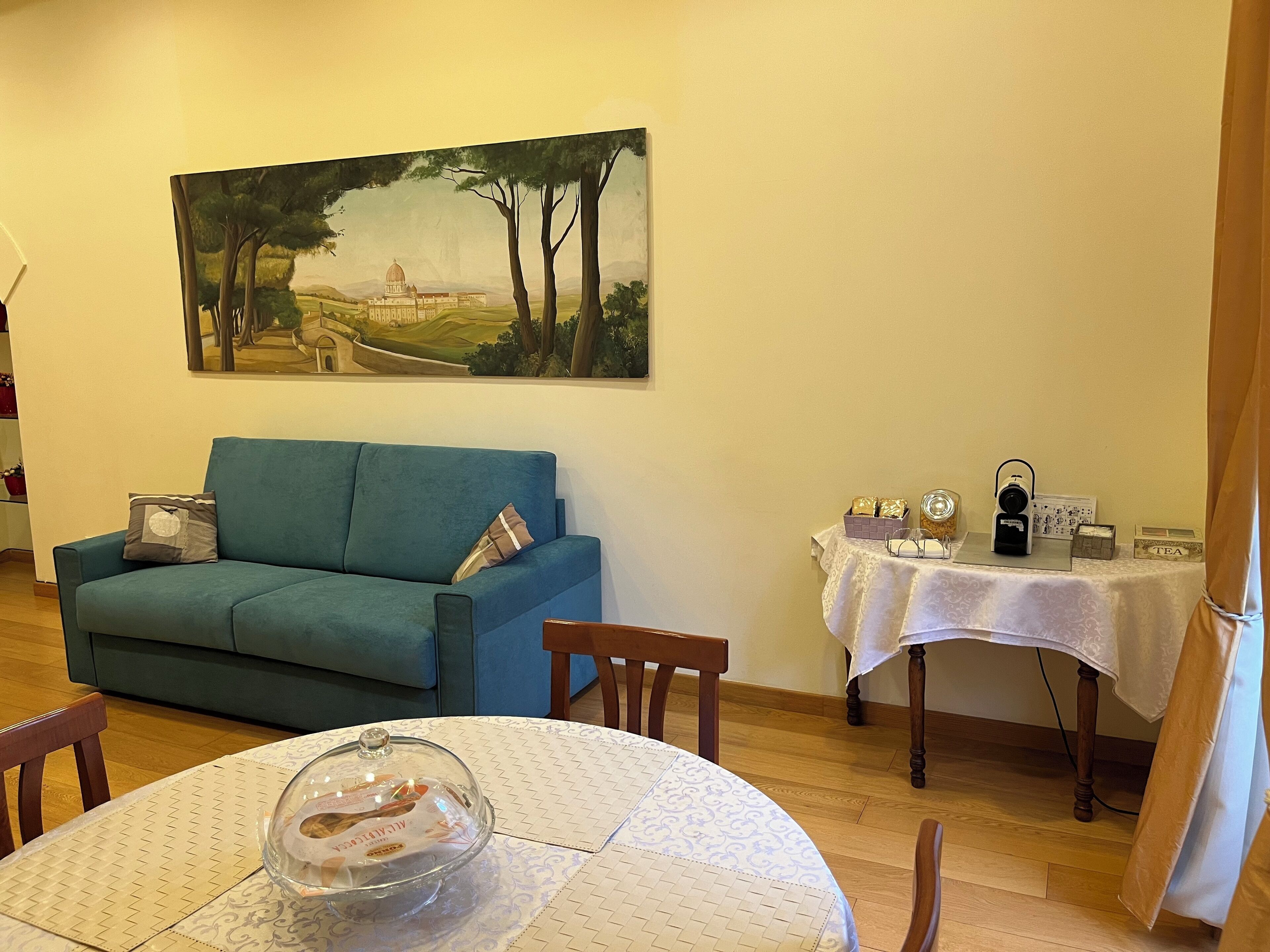 A living room with a blue sofa, a round table with a glass dome centerpiece, a painting of a landscape on the wall, and a small table with tea and a camera on it.