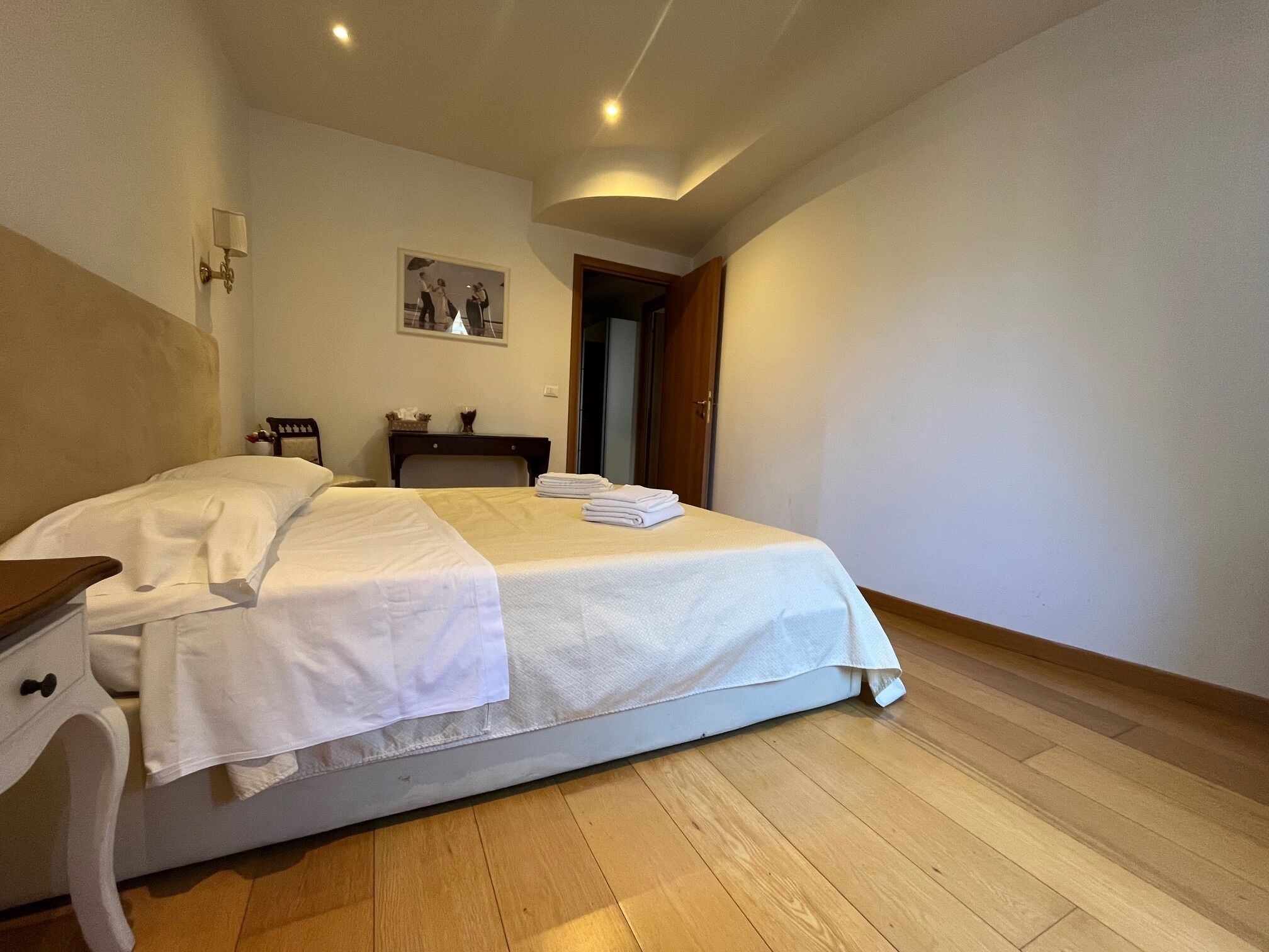 A neatly made bed with white linens, a wooden nightstand with a lamp, and a framed picture on the wall.