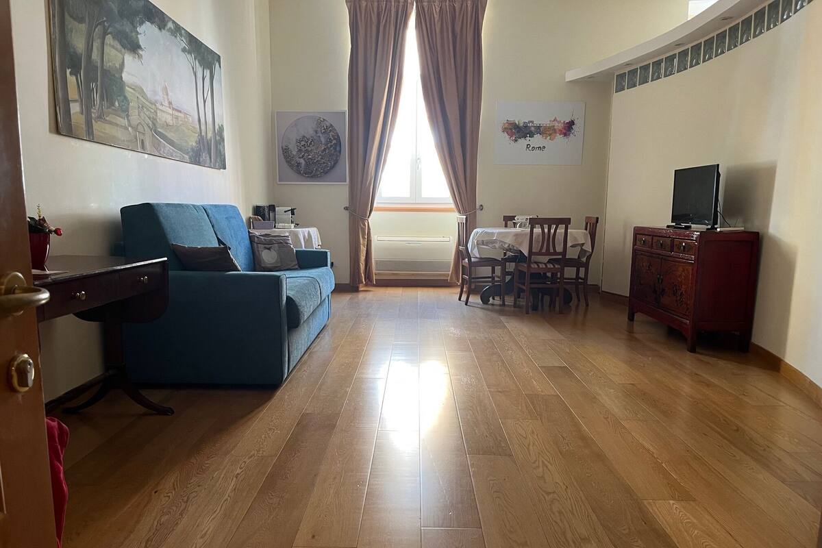 A living room with a blue sofa, a wooden cabinet, a dining table with chairs, and a painting of Rome on the wall.