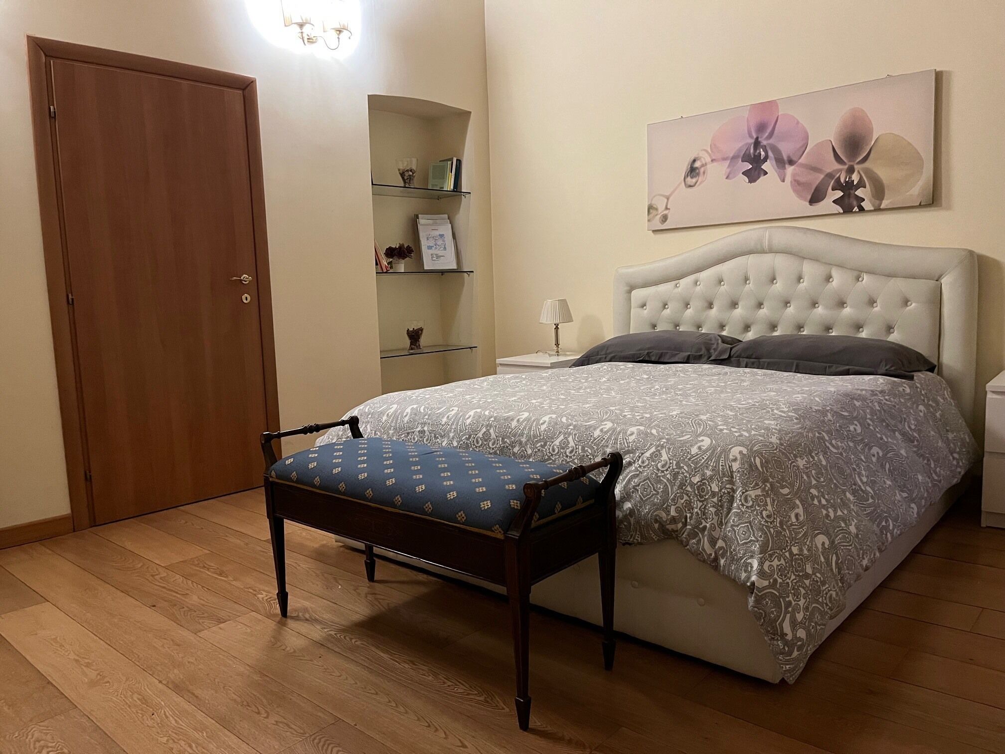 Un dormitorio con cama, mesita de noche, una silla, armario y estante con objetos decorativos.
