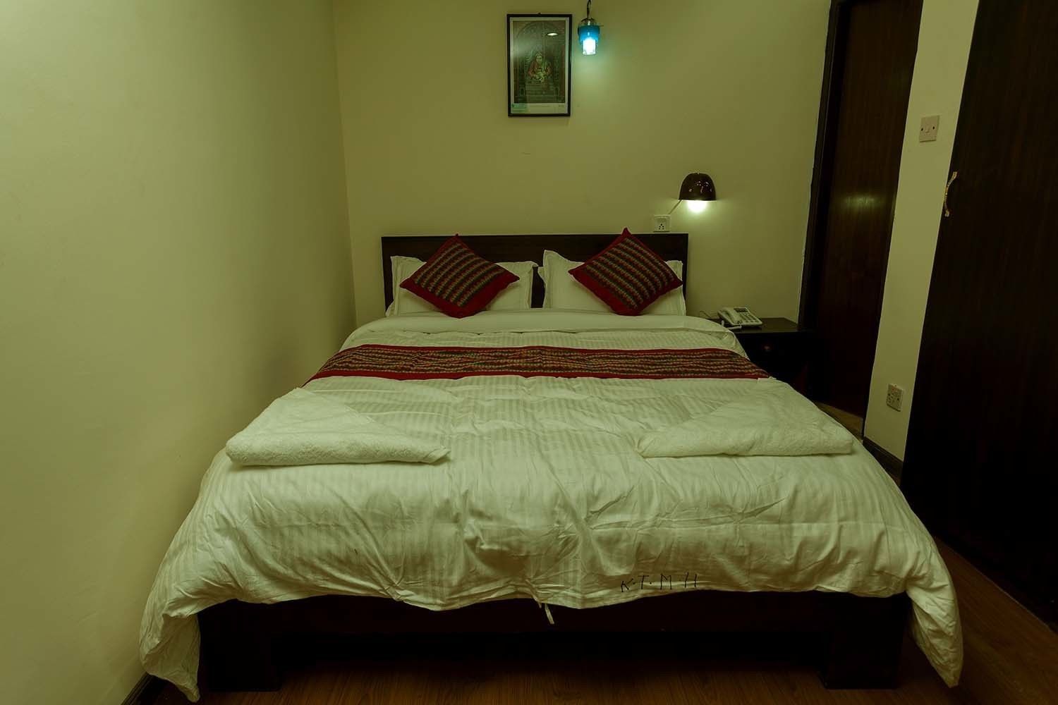 A neatly made bed with a white comforter and red and white pillows, a bedside lamp, and a framed picture on the wall.