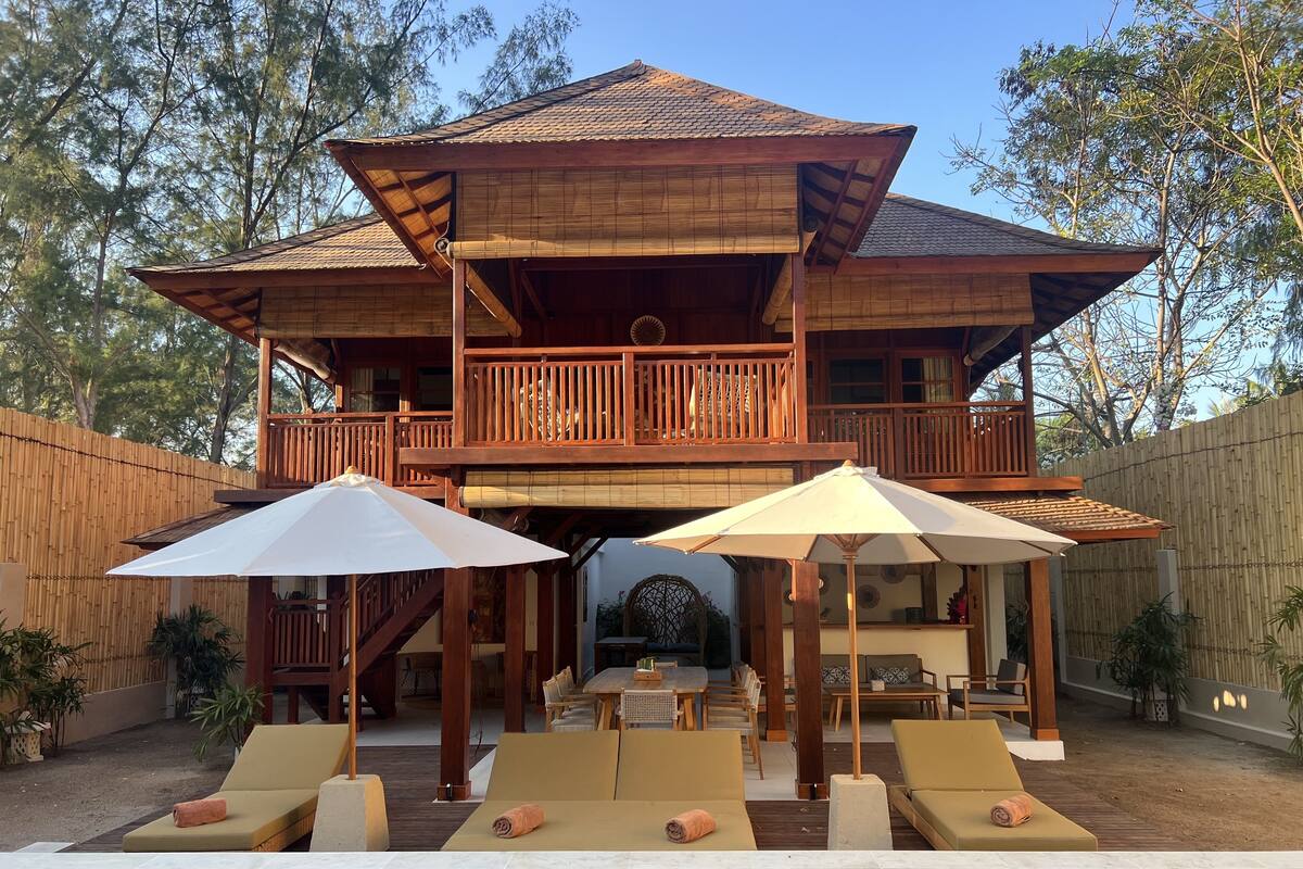 Une maison en bois à deux étages, dotée d’un balcon, d’un espace piscine avec des chaises longues et des parasols, et d’une clôture en bois.