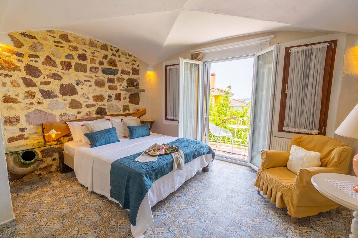 A bedroom with a stone wall, a bed with a blue bedspread, a yellow armchair, and a balcony with a view of greenery.