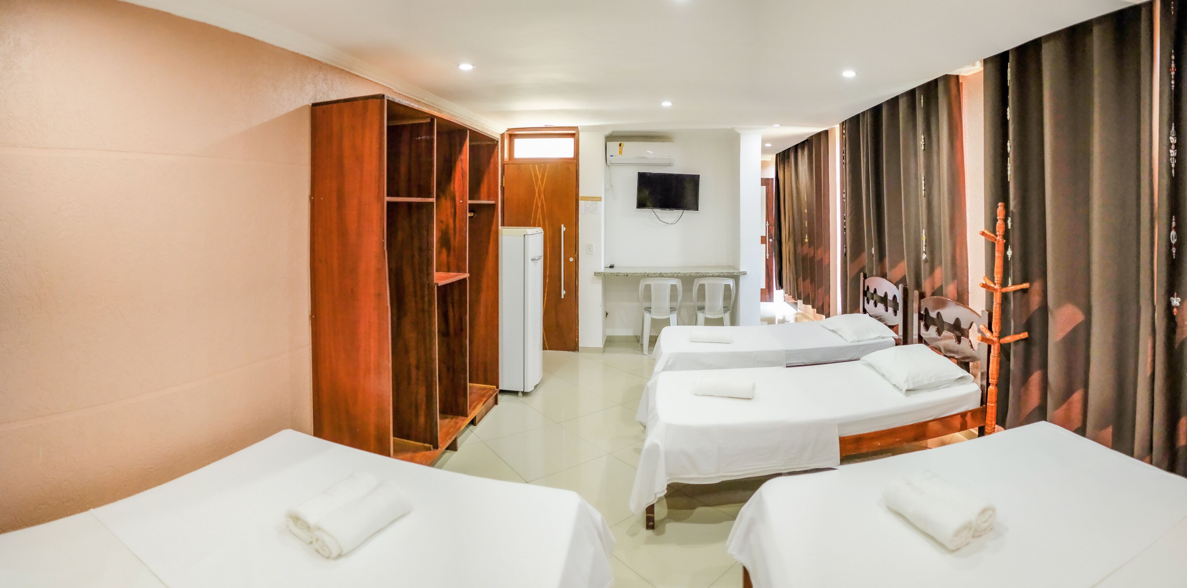 Quarto de hotel com três camas simples, um guarda-roupa, uma televisão e uma pequena área de kitchenette.