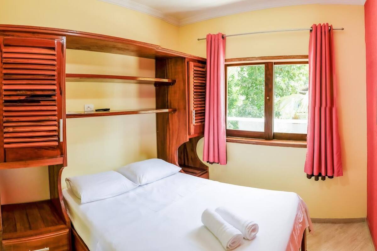 Um quarto com cama, estantes de madeira, janela com cortinas e televisão na parede.