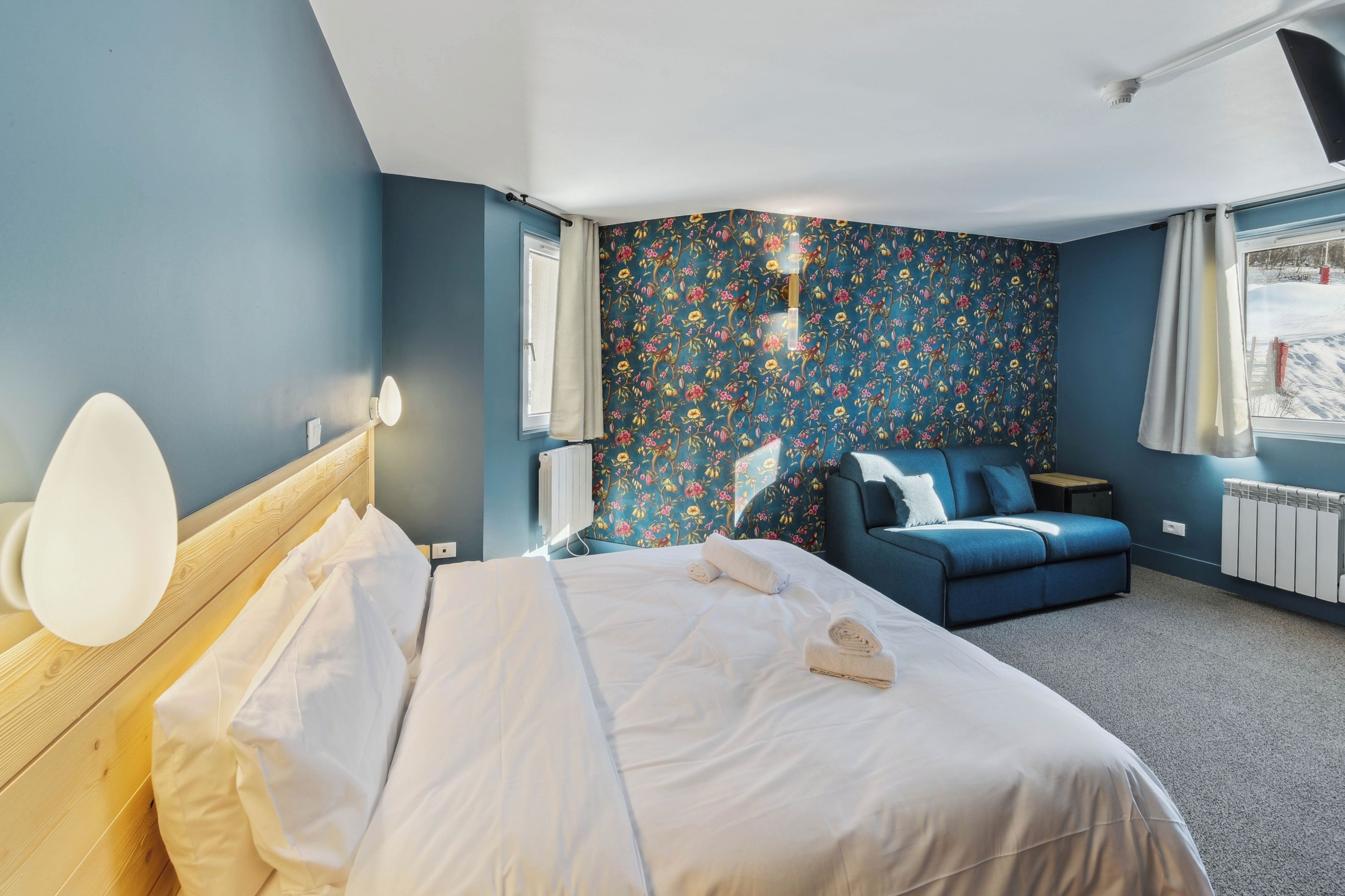 Un dormitorio con una cama, un sofá azul, una ventana con cortinas y una pared con estampado floral.