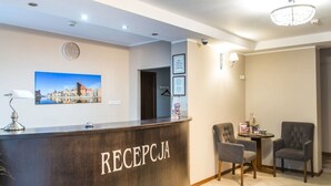 Zona de recepción con un mostrador de madera identificado con la palabra “RECEPCJA”, un reloj, diplomas enmarcados, una pintura de un paisaje urbano y dos sillones.