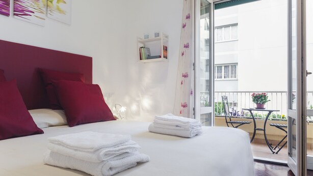 Una habitación de hotel con cama, almohadas rojas, una pequeña estantería con libros y un balcón con mesa y sillas.