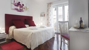 Un dormitorio con cama, mesita de noche, silla, escritorio con una botella y vasos, y ventana con cortinas.