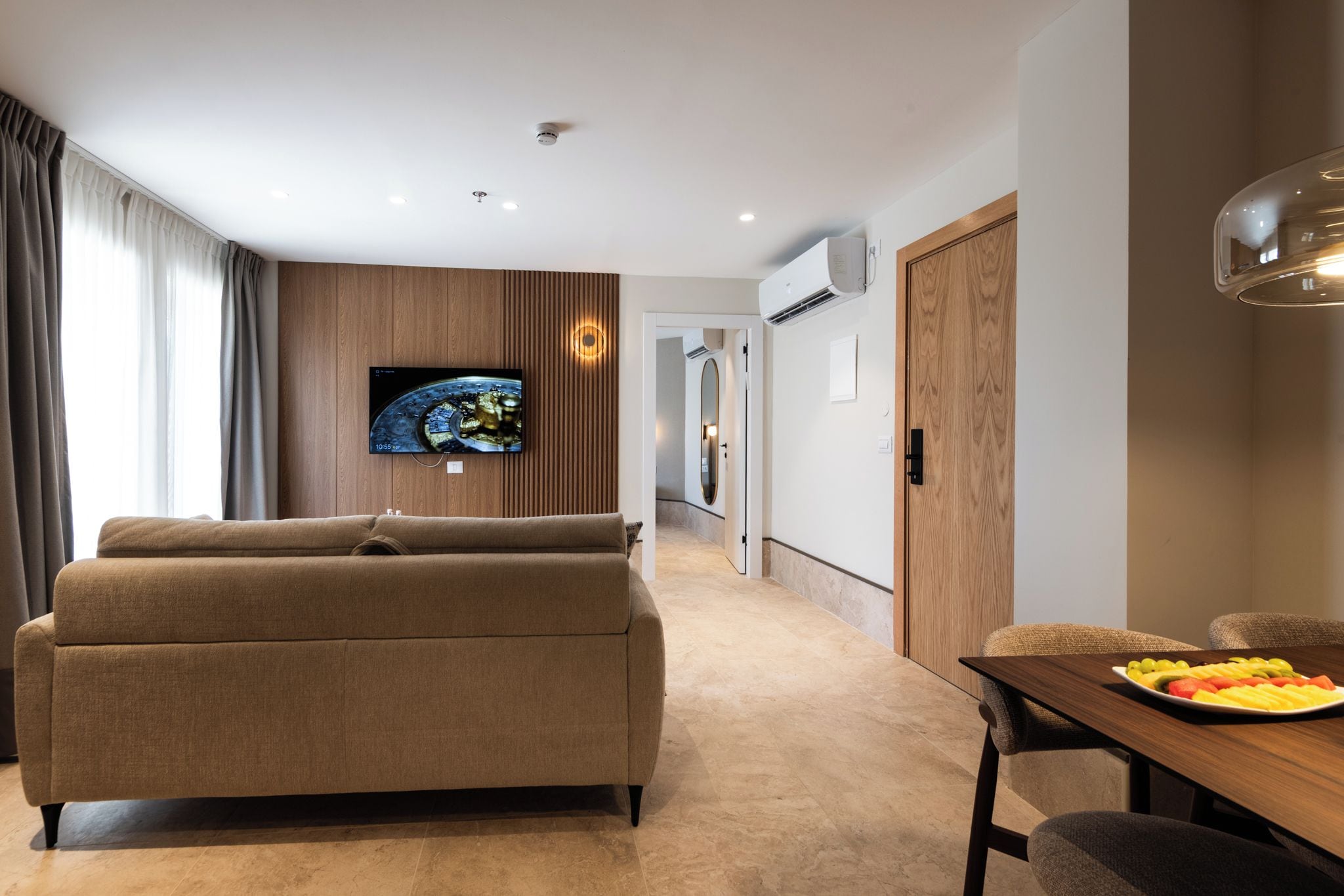 A modern living room with a sofa, a dining table with fruit, a TV mounted on the wall, and a hallway leading to another room.