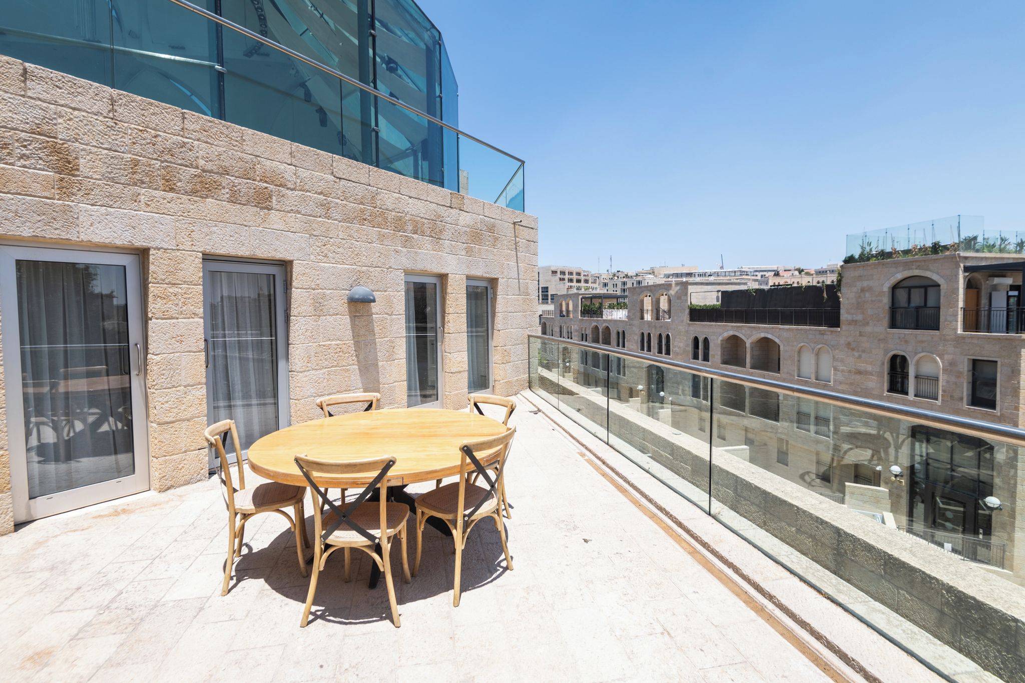 Una terraza en la azotea con una mesa y sillas de madera redonda, barandilla de vidrio y vistas panorámicas de la ciudad.