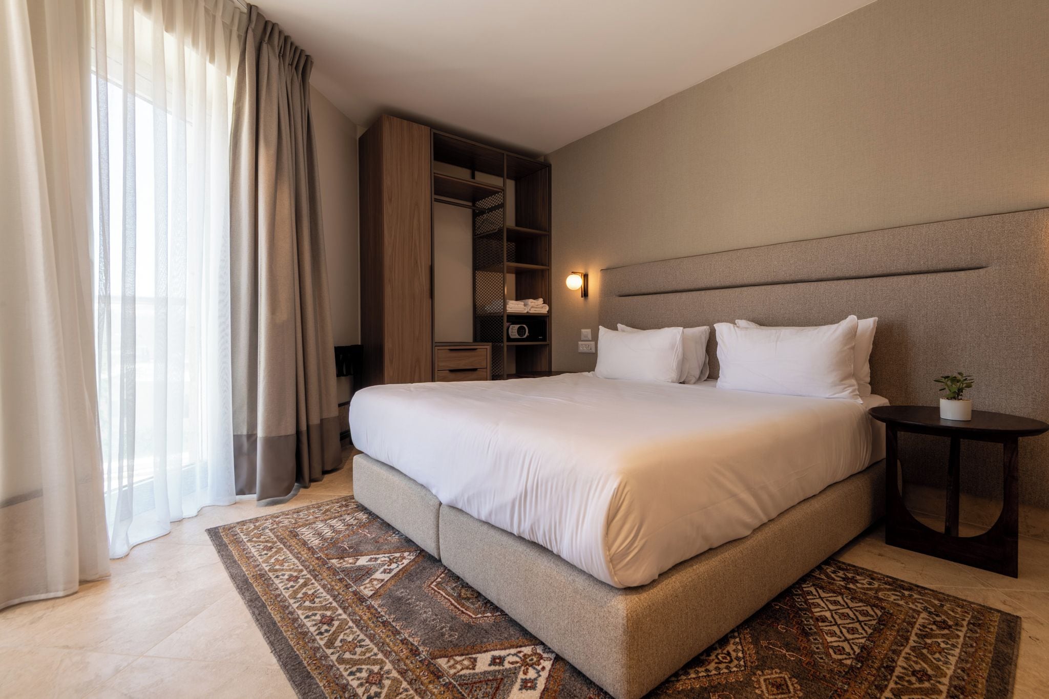 A hotel room with a large bed, a bedside table, a nightstand, a built-in wardrobe, and a large window with curtains.
