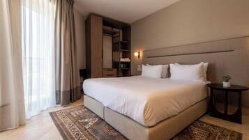 A hotel room with a large bed, a bedside table, a nightstand, a built-in wardrobe, and a large window with curtains.