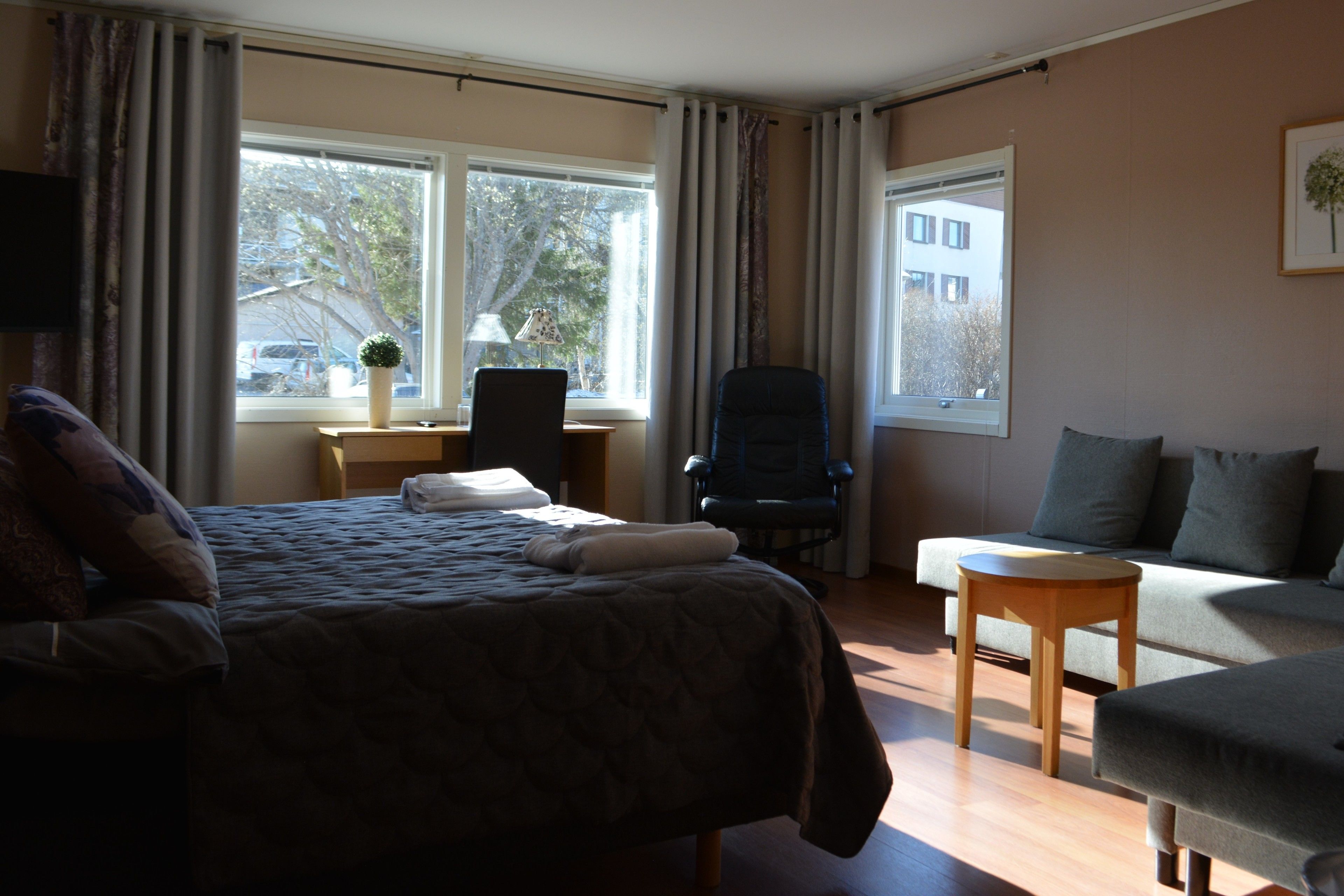 Quarto de hotel com cama, escrivaninha com luminária, poltrona, sofá e janela com cortinas.