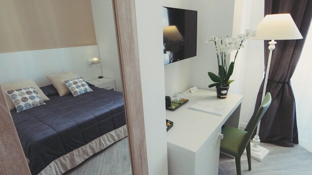 A modern bedroom with a bed, a desk, a chair, a mirror, a lamp, and a potted plant. - Vantaggio Suites (Rome)