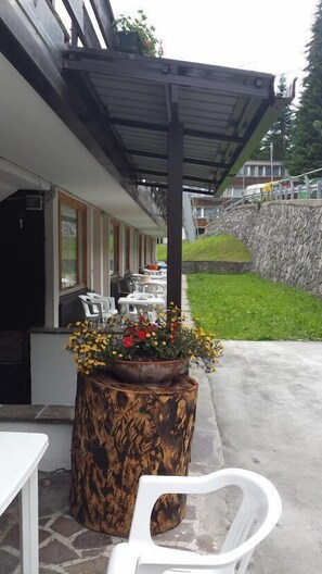 An outdoor patio with a covered seating area, a large planter with flowers, and a stone wall in the background. - TH 1400 (Mezzana)