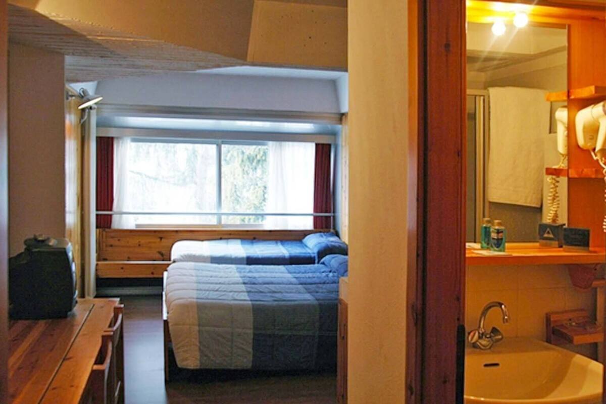 Une chambre à coucher de taille modeste, comprenant un lit, une fenêtre et une salle de bain avec un lavabo et un miroir.
