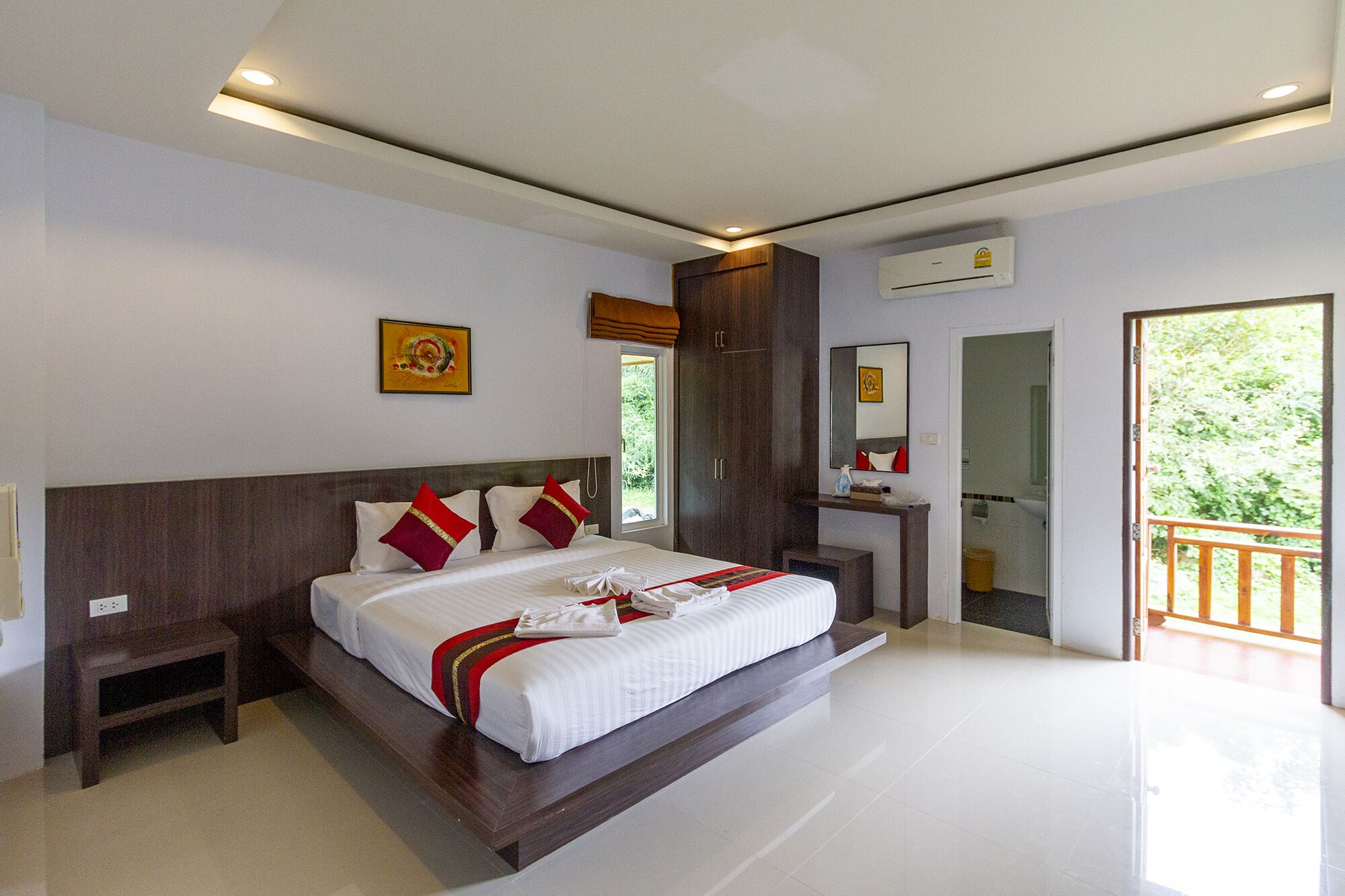 A modern hotel room with a large bed, a nightstand, a wooden wardrobe, a bathroom with a mirror, and a balcony with a view of greenery.