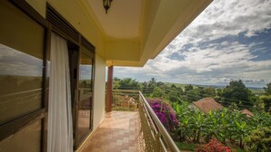 A balcony with a tiled floor, a glass door with curtains, and a view of a garden with banana trees and purple flowers.