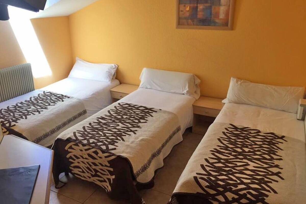 A hotel room with three single beds, each with a patterned bedspread. There is a painting on the wall above the beds and a window providing natural light.