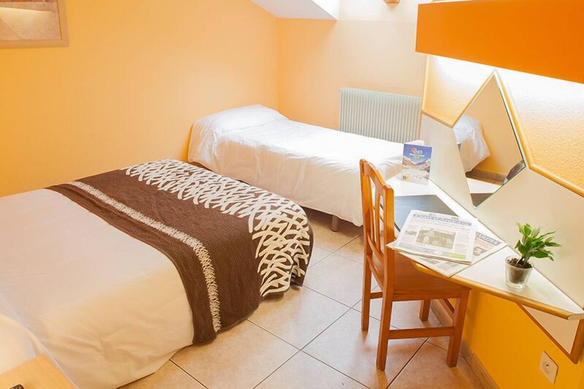 A small, bright room with two beds, a desk, and a chair.