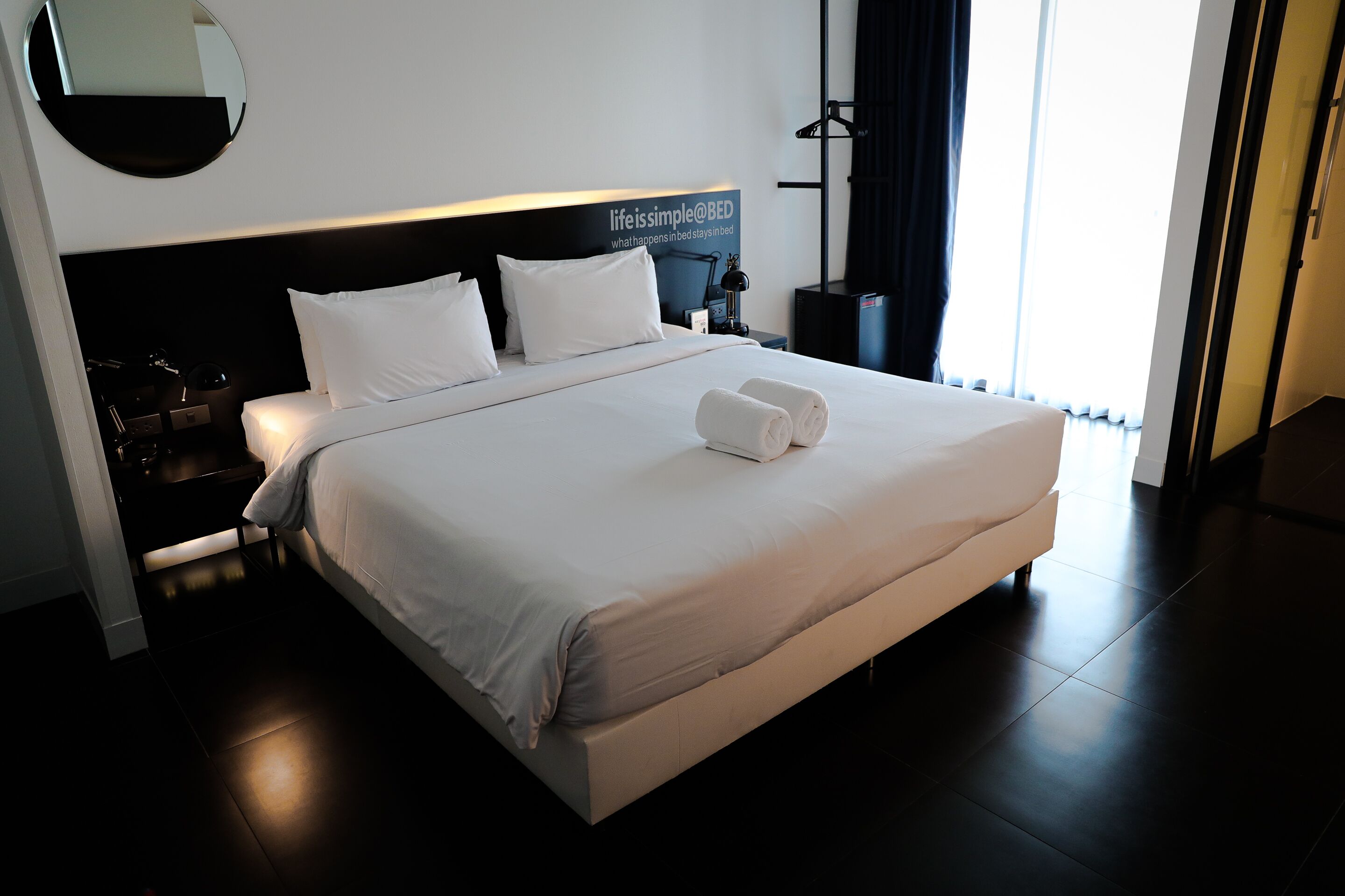 A neatly made bed with white linens and two rolled towels, a bedside table with a lamp, and a wall-mounted mirror.