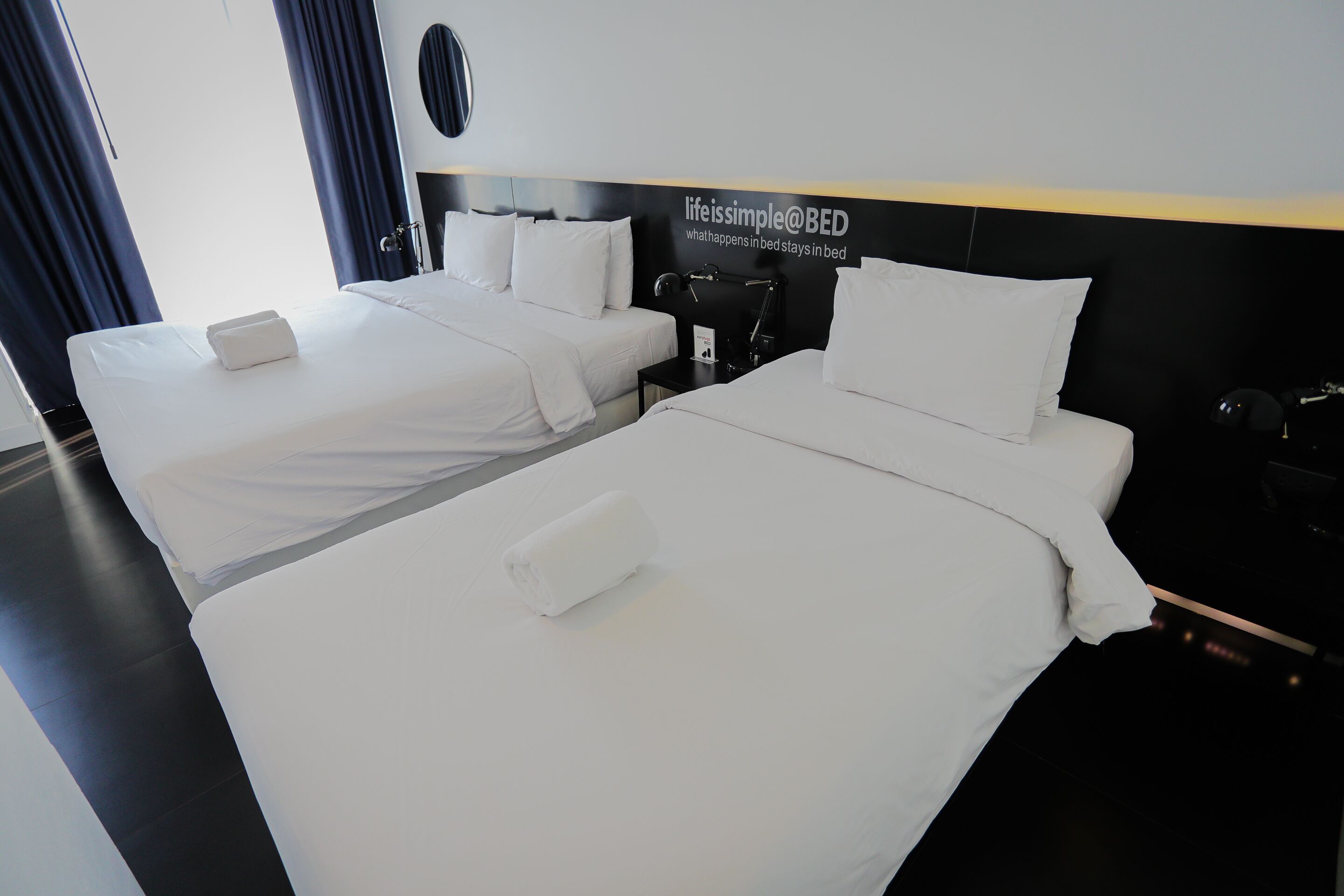 A hotel room with two beds, a black headboard, and a sign reading "life is simple @ BED".