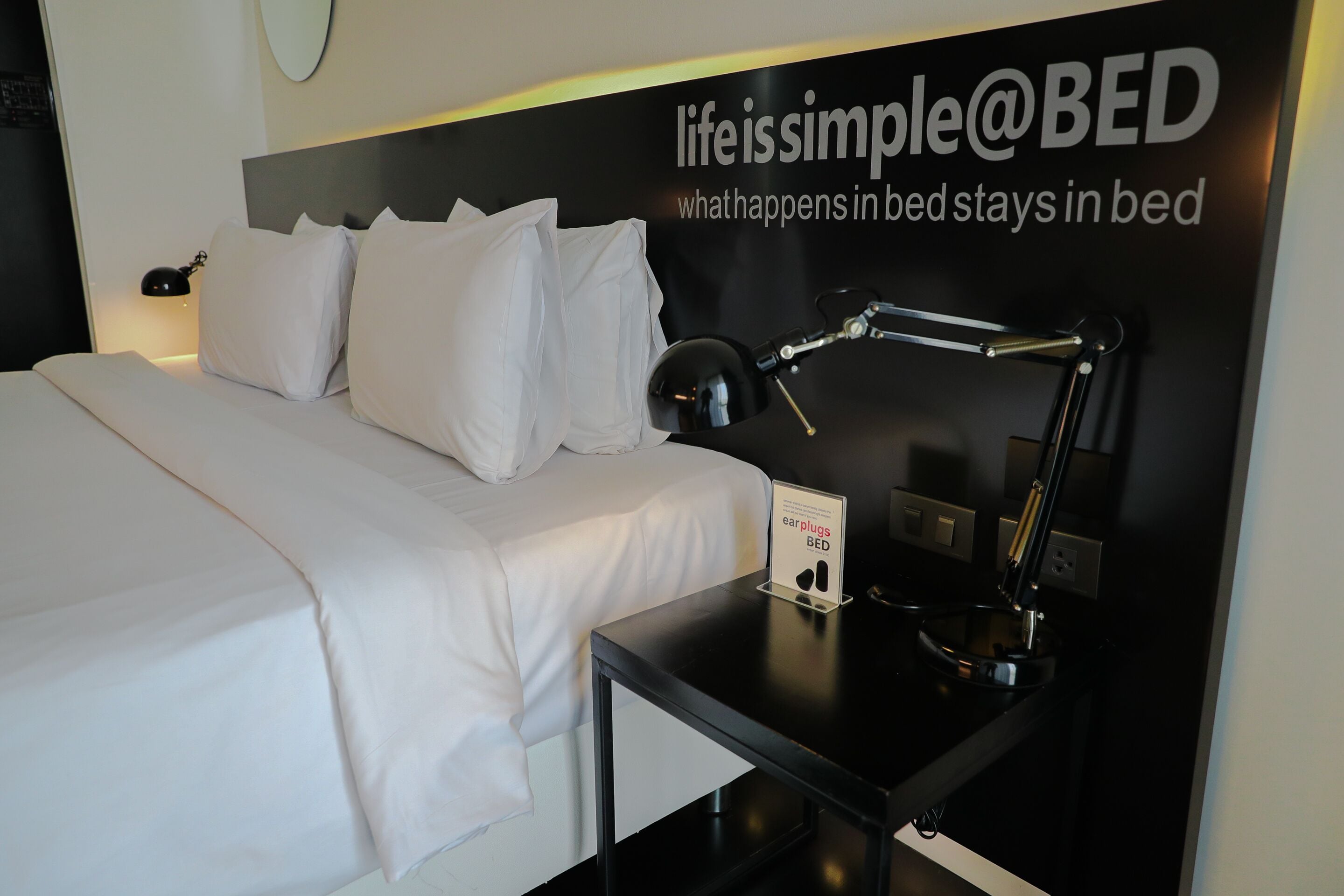 A neatly made bed with white linens, a black bedside table with a lamp, and a sign advertising earplugs.