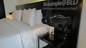 A neatly made bed with white linens, a black bedside table with a lamp, and a sign advertising earplugs.
