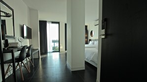Una habitación de hotel moderna con una cama, un escritorio con sillas, un televisor y un ventanal amplio.