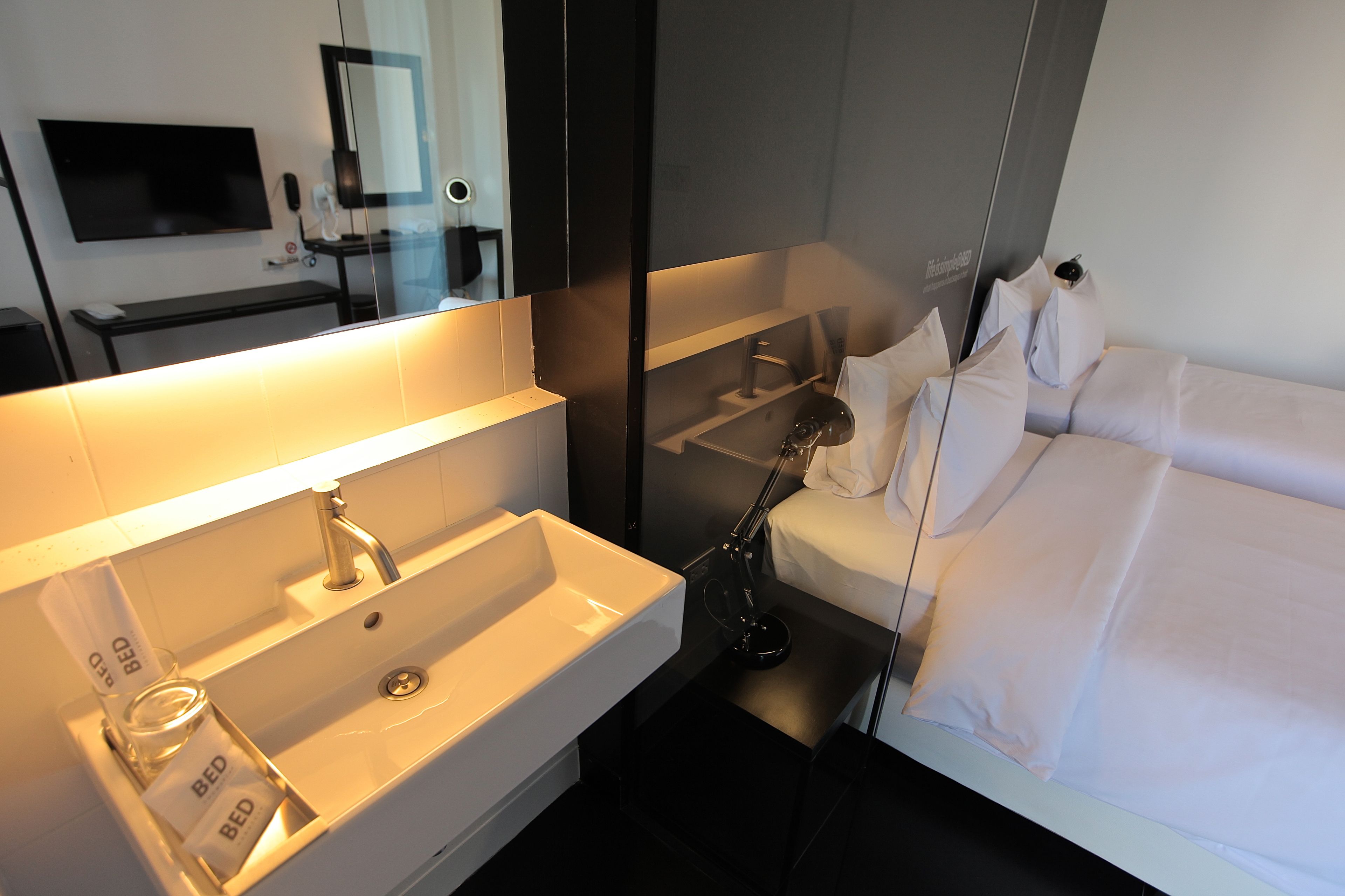 A modern hotel bathroom with a large mirror, a sink, and a bed.