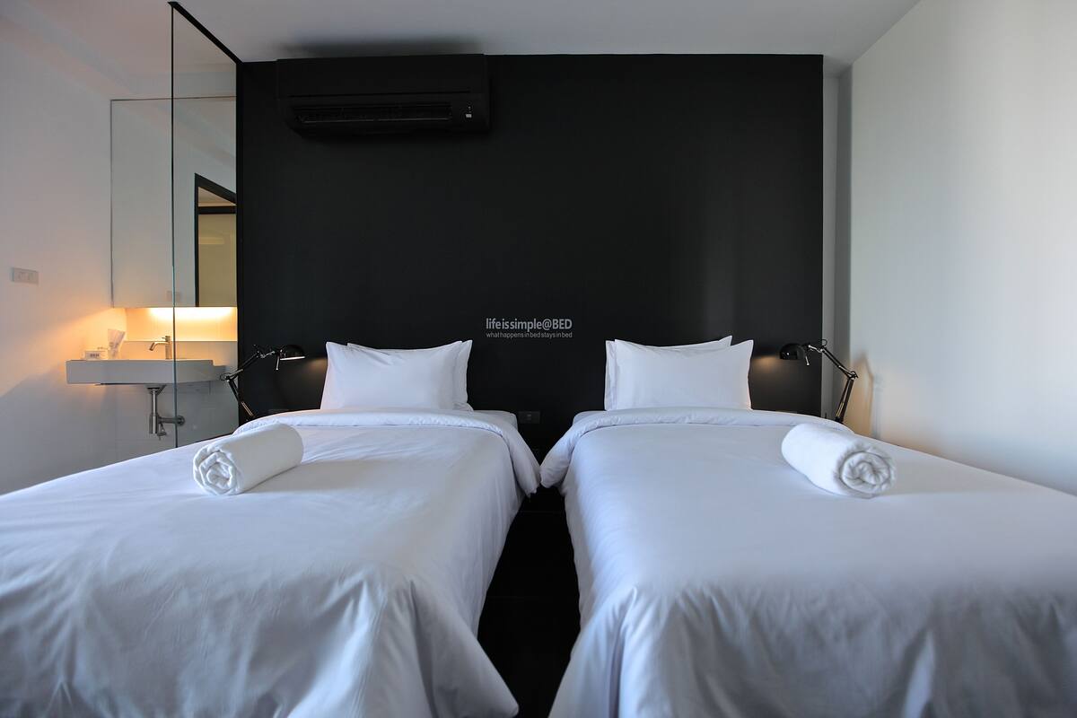 Une chambre d’hôtel avec deux lits simples, une table de chevet avec une lampe, un miroir et un climatiseur fixé au mur.