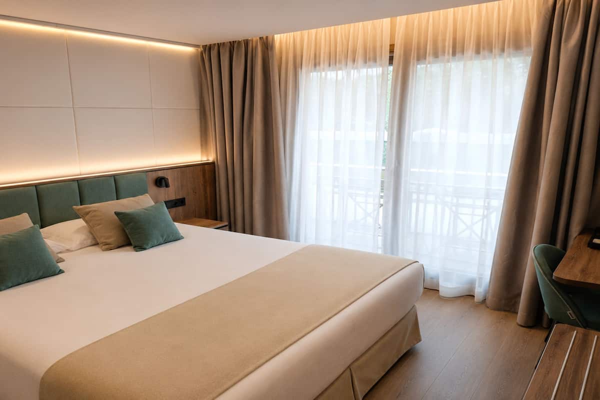 Habitación de hotel con una cama grande, cabecera de madera, mesita de noche y vistas al exterior a través de cortinas traslúcidas.
