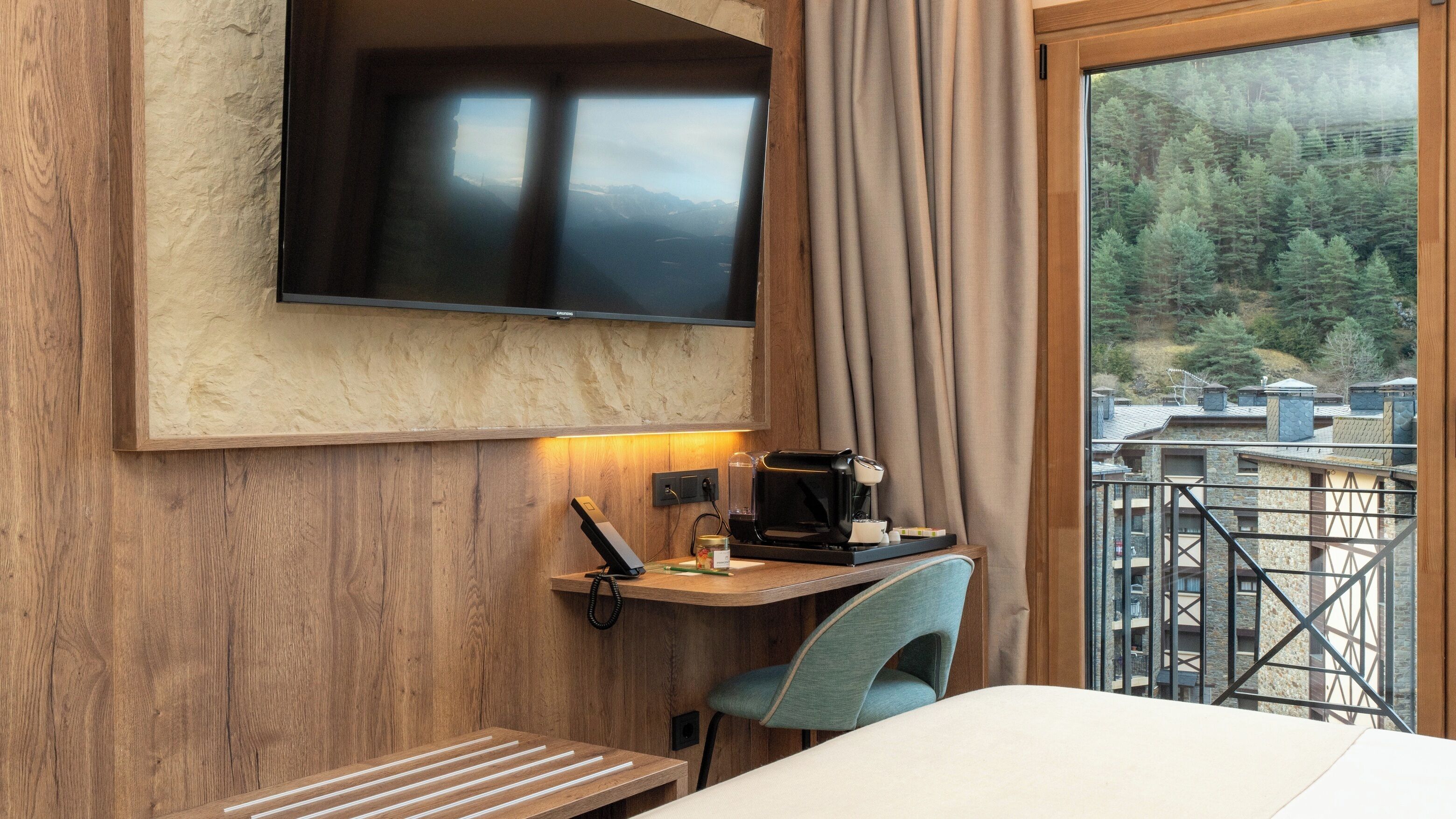 A hotel room with a wooden wall, a flat-screen TV mounted above a desk, a chair, a bed, and a balcony with a view of a forested area.