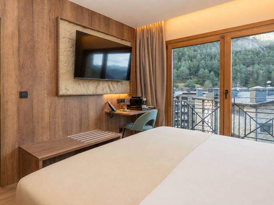 A hotel room with a bed, a desk, a TV, and a balcony with a view of trees.