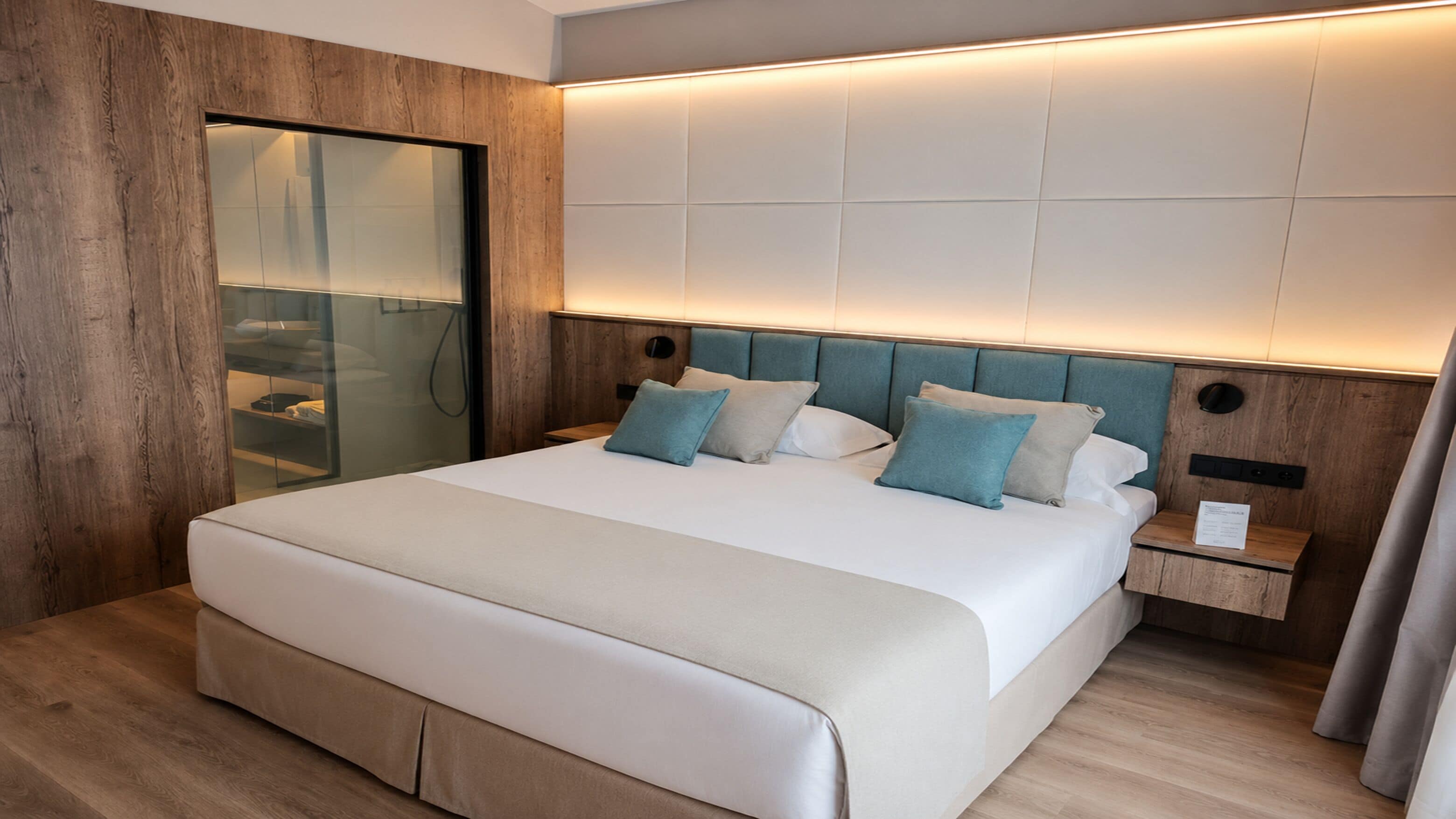 Une chambre d’hôtel moderne avec un grand lit, des panneaux de bois, une douche en verre et une table de chevet avec une lampe.