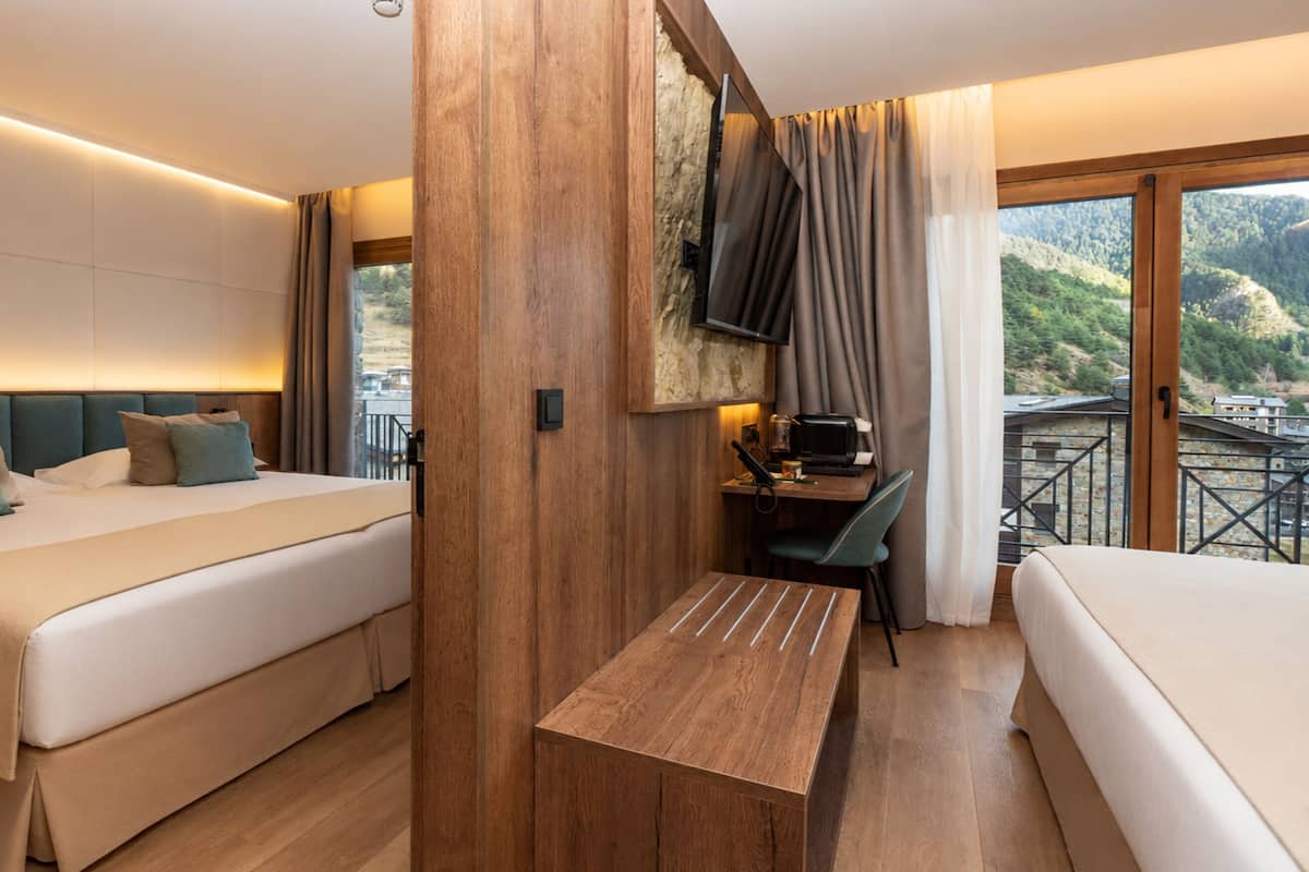 Habitación de hotel con una cama grande, un escritorio con una silla, un banco de madera y vistas a un paisaje montañoso.