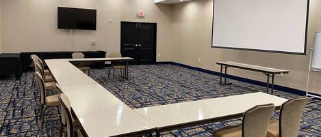 A conference room with a long table, chairs, a projector screen, and a flat-screen TV.