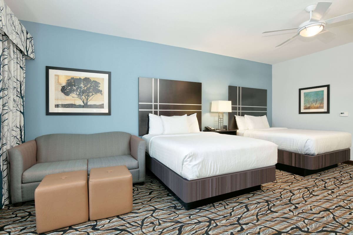 A hotel room with a sofa, two beds, a ceiling fan, and a patterned carpet.