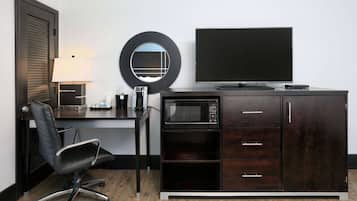A hotel room with a dark wood desk, a black office chair, a flat-screen TV, a microwave, a coffee maker, and a round mirror.