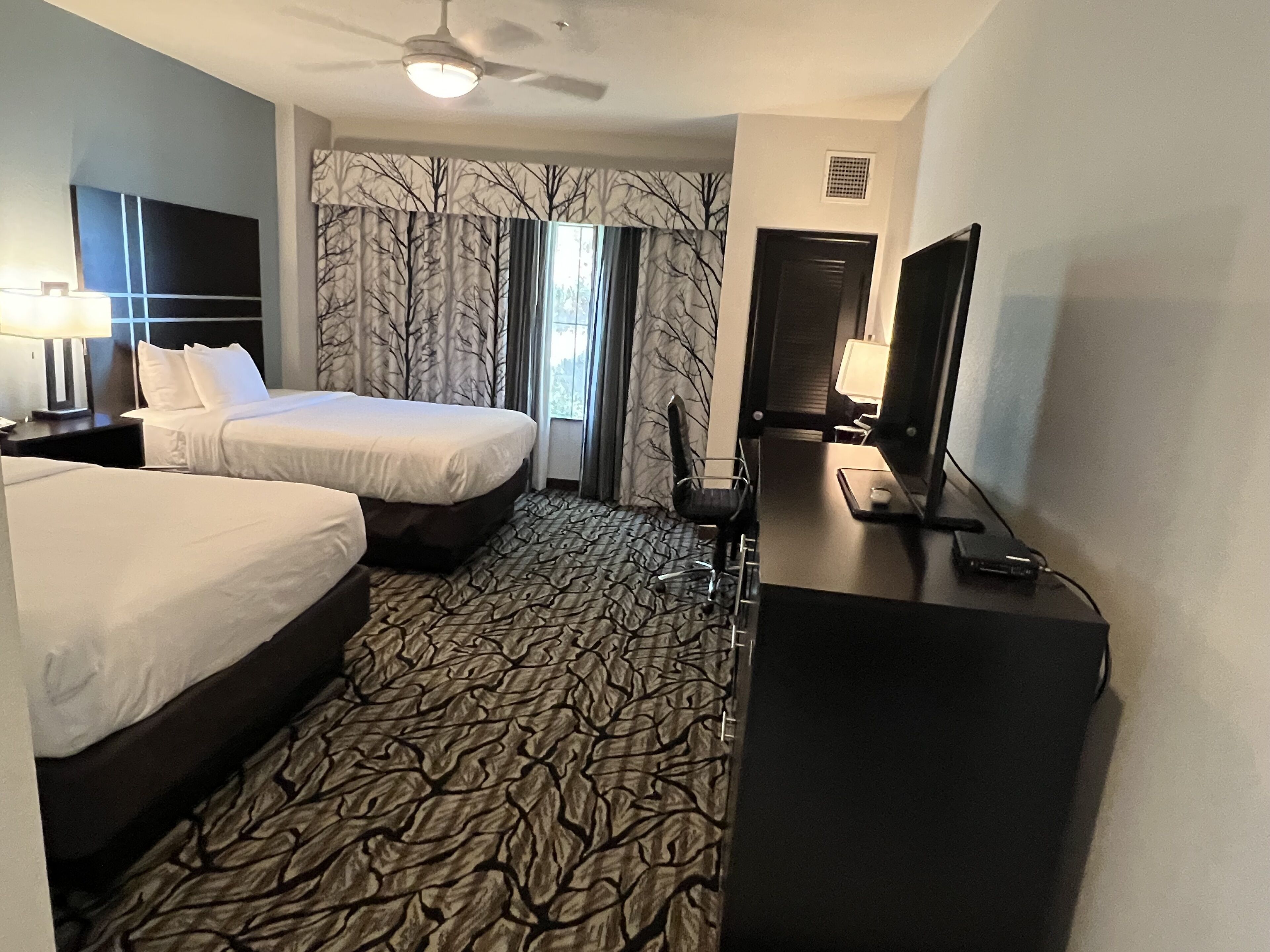 A hotel room with two beds, a desk, a chair, a television, and a ceiling fan.
