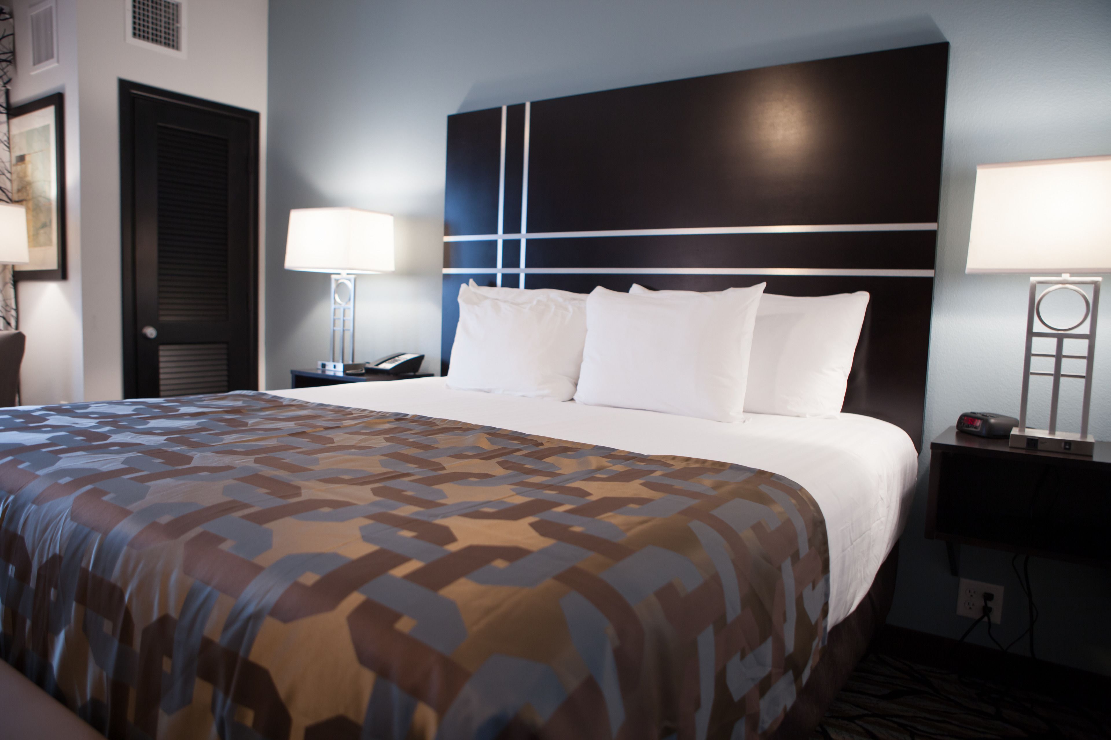 A hotel room with a large bed, a headboard with a geometric design, bedside lamps, and a nightstand with a phone and a clock.