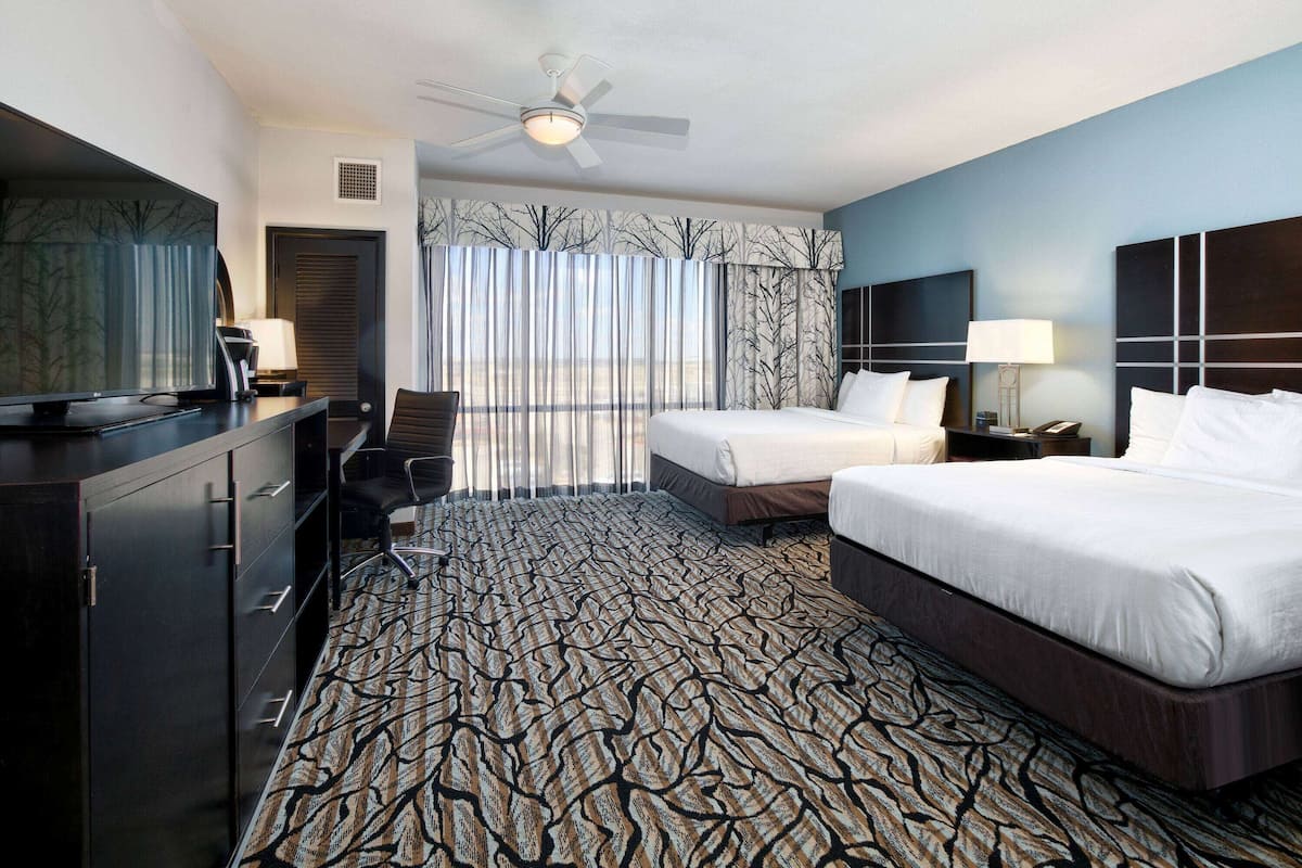 A hotel room with a large bed, a smaller bed, a desk with a chair, a television, and a ceiling fan.