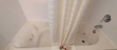 Una bañera blanca con cortina de ducha, una cesta pequeña con artículos de aseo y una toalla en el suelo.