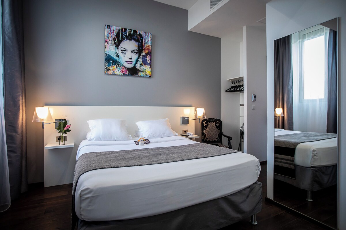 A hotel room with a large bed, bedside lamps, a mirror, a painting on the wall, and a small table with a vase and flowers.