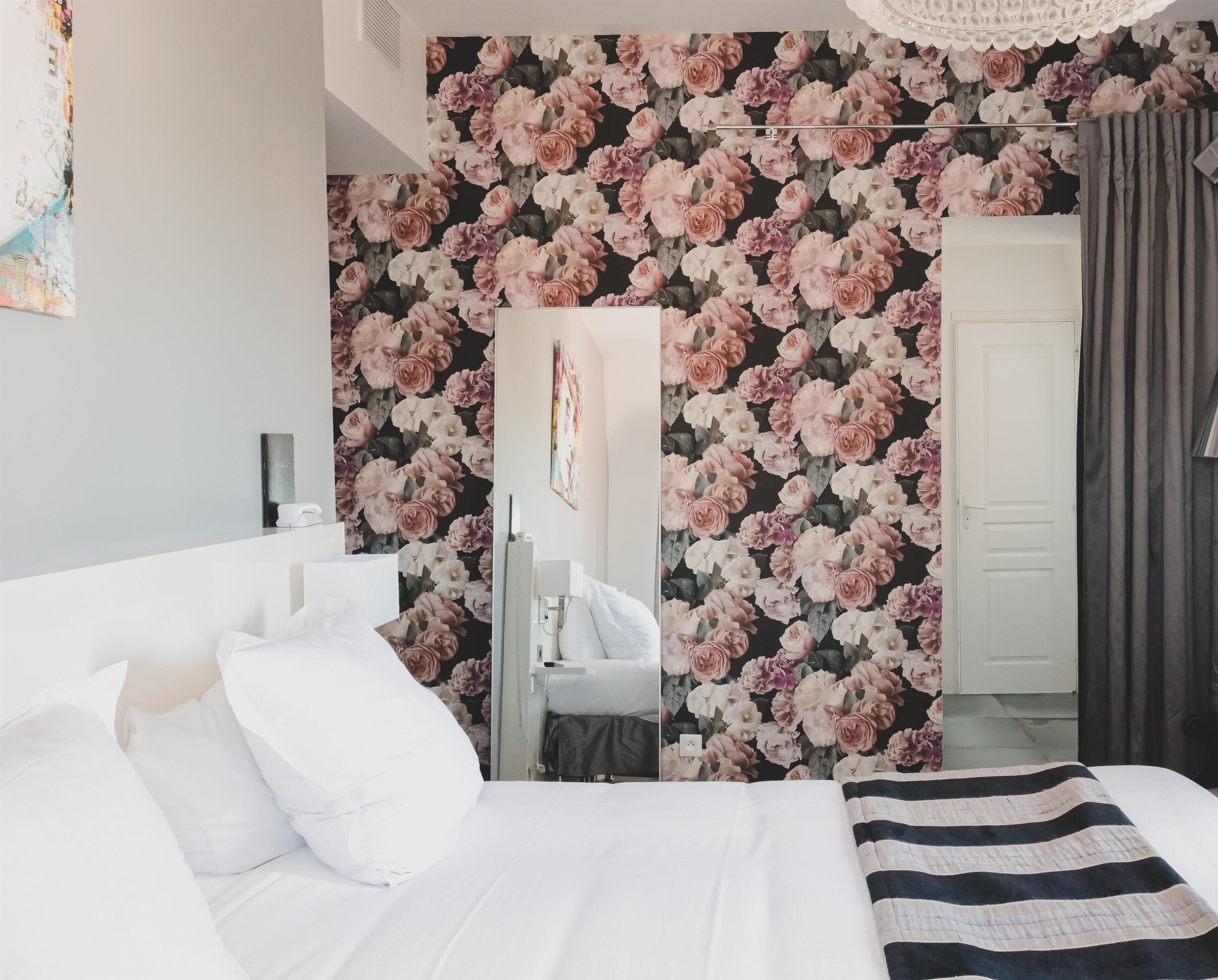 Une chambre à coucher avec un lit, des oreillers blancs, une couverture à rayures, un miroir et un papier peint à motifs floraux.