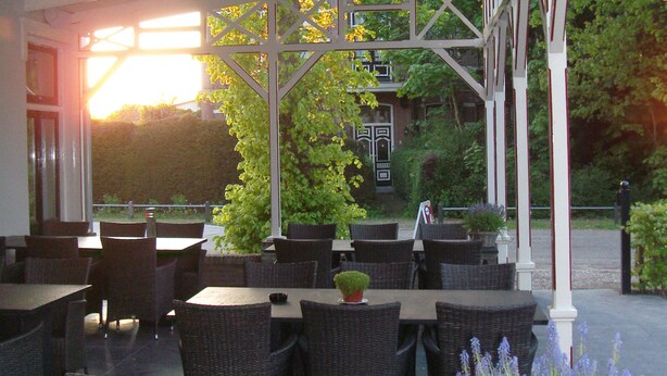 An outdoor cafe with wicker tables and chairs, a potted lavender plant, and a view of a building with trees in the background. - Hotel Restaurant het Rechthuis (Muiderberg)